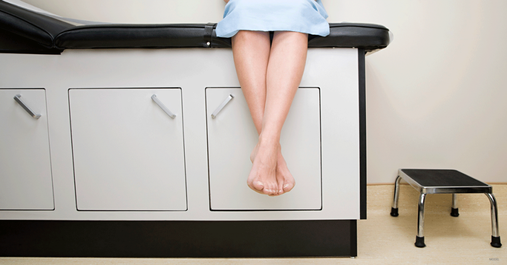 Woman sitting on exam table in surgical gown with legs crossed at the ankle