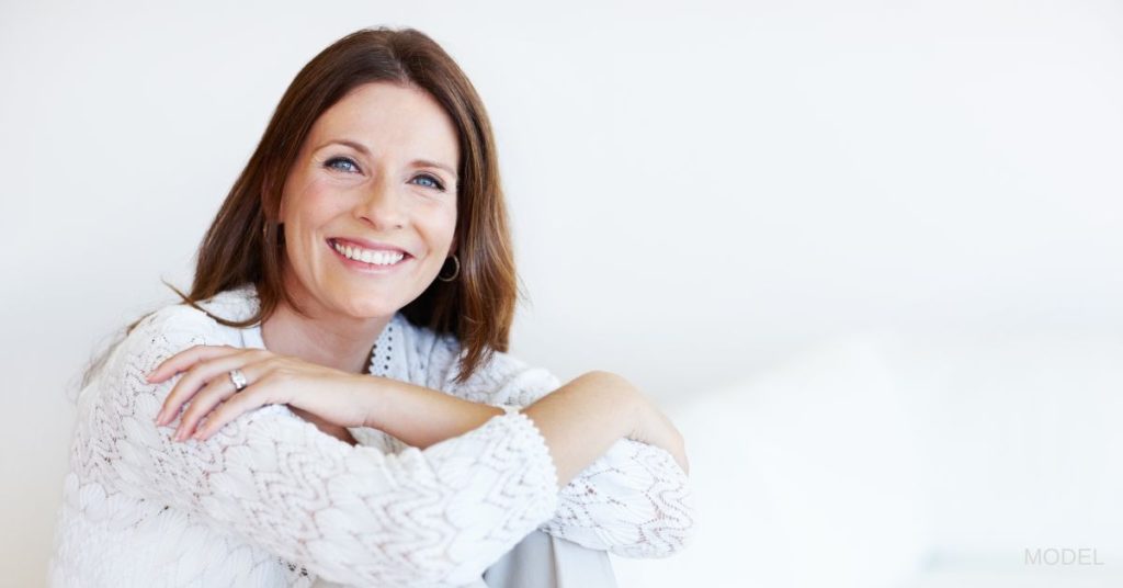 A beautiful middle-aged woman in a white sweater, hugs herself while smiling at the camera. (Model)